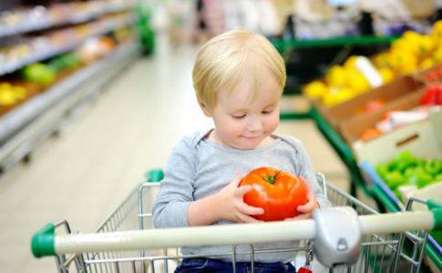 Kleines Kind, das im Einkaufswagen sitzt und eine Tomate in den Haenden haelt