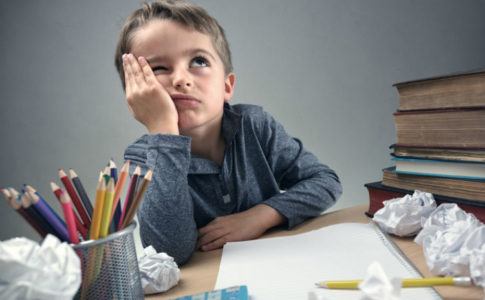 Kind sitzt am Schreibtisch zwischen Büchern und zusammengeknülltem Papier und kämpft mit Hausaufgaben