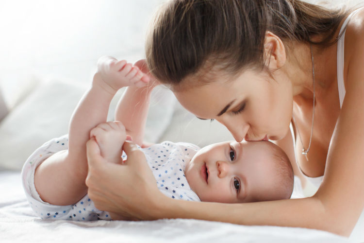 Mutter gibt ihrem lachendem Baby im Strampler einen Kuss auf den Kopf