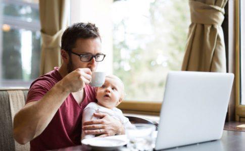 Selbstständiger Mann sitzt mit Baby vor dem Laptop und arbeitet