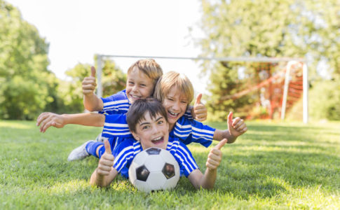 Drei Jungs liegen draußen mit einem Fußball auf der Wiese und lachen
