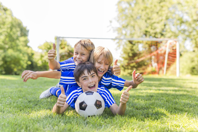 Drei Jungs liegen draußen mit einem Fußball auf der Wiese und lachen