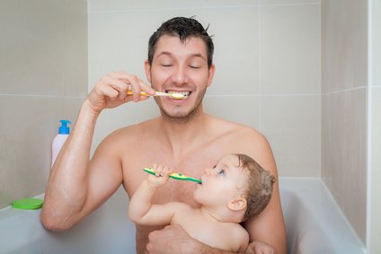 Ein Vater sitzt mit seinem Kind in der Badewanne und beide putzen Zähne.