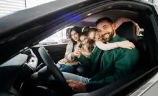 Familie im Auto, Vater hält Autoschlüssel, Kinder umarmen ihn auf dem Fahrersitz.