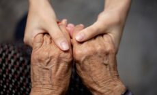Zwei Menschen halten sich gegenseitig an den Händen. Eine Person älter, die andere deutlich jünger.
