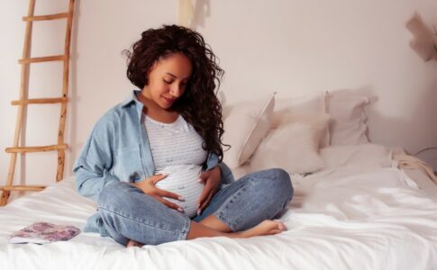 Eine schwangere Frau sitzt auf dem Bett und fasst sich an den Bauch