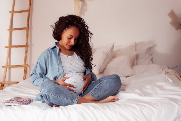 Eine schwangere Frau sitzt auf dem Bett und fasst sich an den Bauch