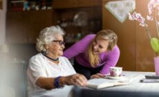 Eine ältere Dame sitzt an einem Tisch. Eine Pflegekraft steht neben ihr und lächelt.