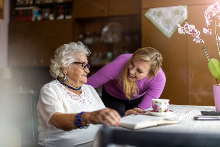 Eine ältere Dame sitzt an einem Tisch. Eine Pflegekraft steht neben ihr und lächelt.