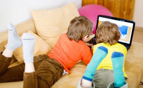 Zwei Kinder liegen auf dem Sofa und gucken auf einen Laptop Bildschirm