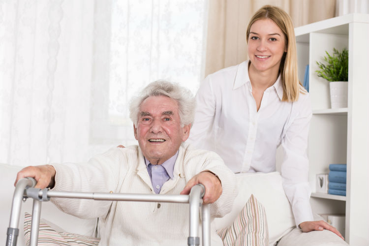 wohnen im alter - alter Mann situt mit Gehhilfe vor sich auf Sofa & junge Frau steht hinter ihm